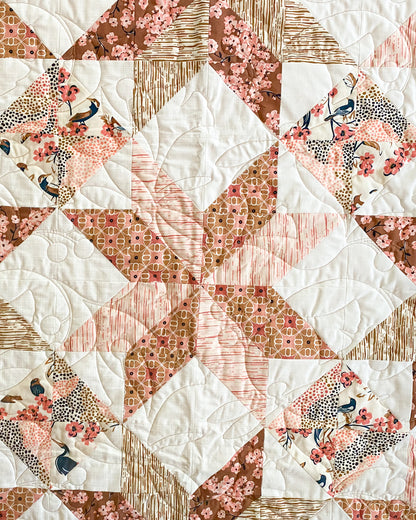 A close-up of a quilted textile featuring a geometric starburst pattern in white, blush pink, peach, and brown, with floral and bird-print fabric sections.