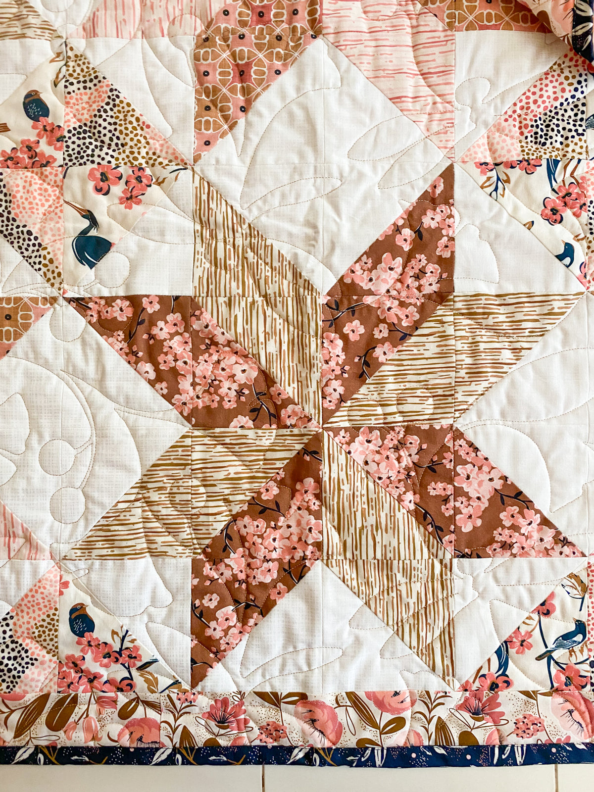 A close-up of a quilted fabric featuring a geometric star and diamond block pattern in white, brown, pink, and floral prints with bird motifs.