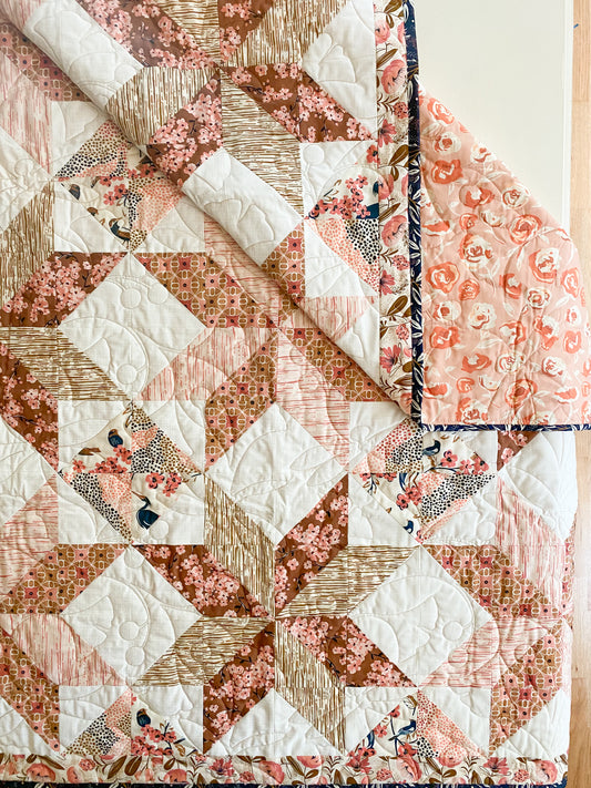 A quilted patchwork quilt shown laid flat on a light wood surface, with one corner folded back to reveal the reverse side. The design combines cream, blush pink, peach, brown, and small floral/animal-print sections in a geometric star and diamond pattern.