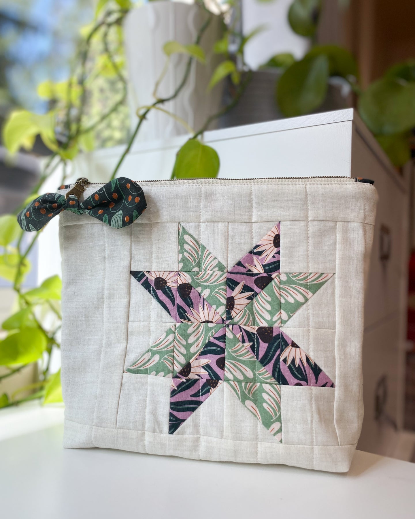 Zipper pouch made from PDF sewing pattern, featuring purple and green Cloud9 organic floral fabric and flat-bottom design, standing on a white table with greenery behind